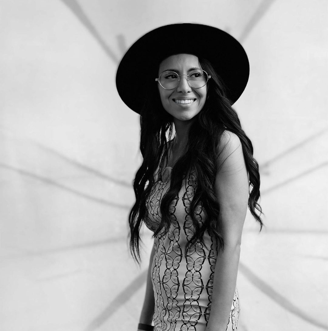 Black and white portrait of Sarai Mora in a wide-brimmed hat, smiling