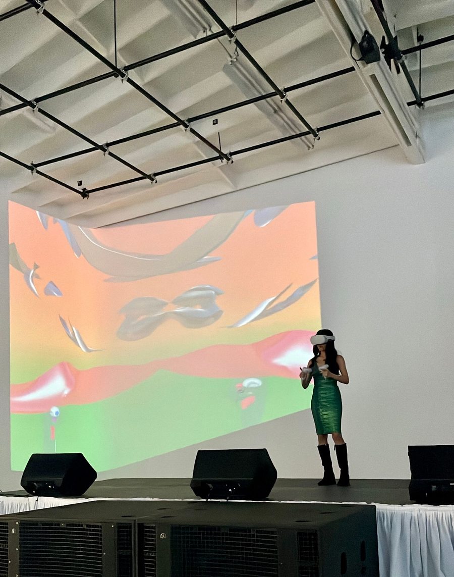 Creatress on a gallery stage in front of a wall-sized projection of her sculpture