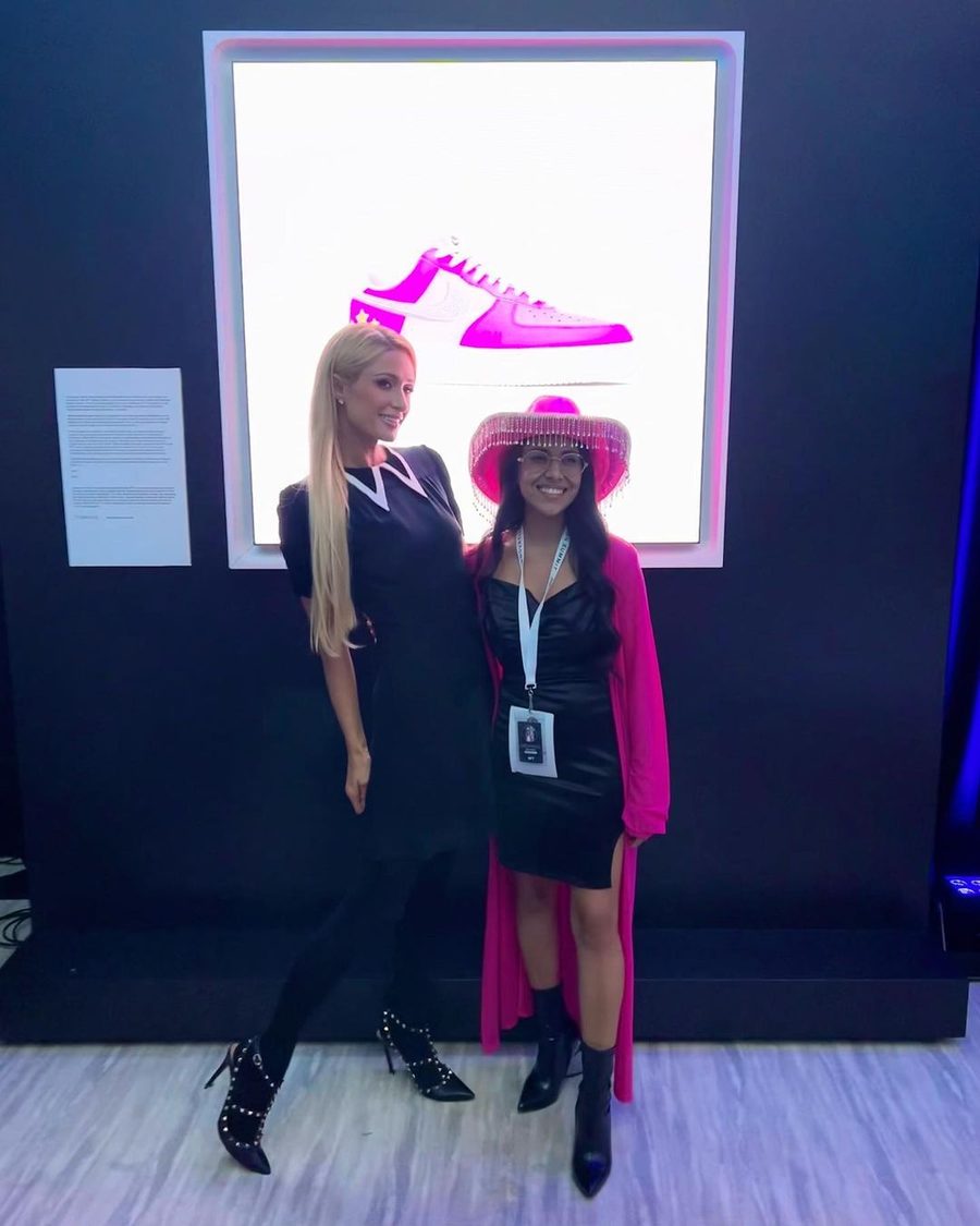 Creatress in a pink fringed hat standing with Paris Hilton in front of a lit artwork